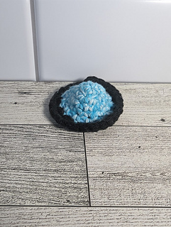 A crochet fidget with a purple center and a light blue rim. The shape of the fidget resembles a sombrero. The background is a light wood grain and a white tile.