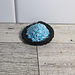 A crochet fidget with a purple center and a light blue rim. The shape of the fidget resembles a sombrero. The background is a light wood grain and a white tile.