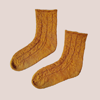 Mustard-gold socks with a knotty-cable pattern on a garter background with a baby hem cuff. They are on a dusty pink background.