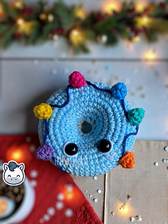 Crochet amigurumi pattern of two Christmas donuts: one beige with white icing and holly, and one blue with a colorful string of Christmas lights