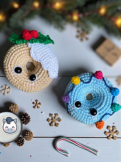 Crochet amigurumi pattern of two Christmas donuts: one beige with white icing and holly, and one blue with a colorful string of Christmas lights