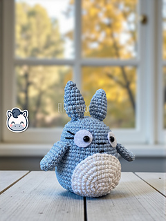 Blue and white crochet Totoro amigurumi on a wooden table with a blurry autumn forest background.