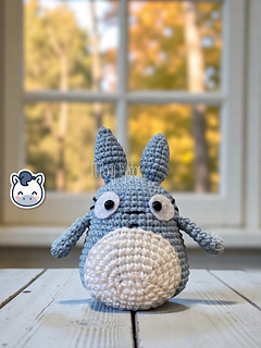 Blue and white crochet Totoro amigurumi on a wooden table with a blurry autumn forest background.