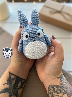 Blue and white crochet Totoro amigurumi on a wooden table with a blurry autumn forest background.