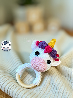 Crochet amigurumi pattern of a baby unicorn rattle with a golden horn, pink and purple mane on a wooden ring.