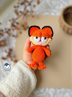 Crochet orange cat amigurumi inspired by Garfield, featuring rounded cheeks, embroidered details and black stripes on the back.