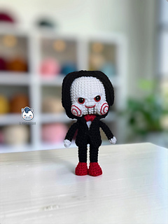 Handmade Billy the Puppet amigurumi inspired by Saw, a horror crochet doll with black suit, red bow tie, red eyes, spiral cheeks and black hair, perfect for Halloween crochet collections.