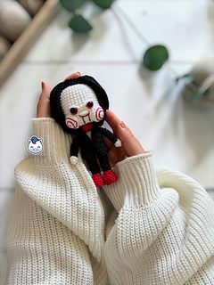 Handmade Billy the Puppet amigurumi inspired by Saw, a horror crochet doll with black suit, red bow tie, red eyes, spiral cheeks and black hair, perfect for Halloween crochet collections.