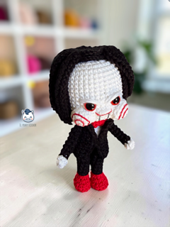 Handmade Billy the Puppet amigurumi inspired by Saw, a horror crochet doll with black suit, red bow tie, red eyes, spiral cheeks and black hair, perfect for Halloween crochet collections.