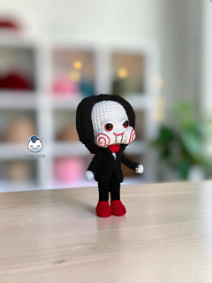 Handmade Billy the Puppet amigurumi inspired by Saw, a horror crochet doll with black suit, red bow tie, red eyes, spiral cheeks and black hair, perfect for Halloween crochet collections.