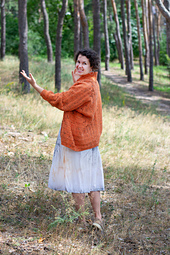 Woman in a cozy crochet cardigan gesturing towards a beautiful forest surrounding her, highlighting the vibrant autumn colors.