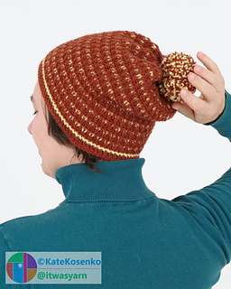 A woman touches the pom-pom on her slouchy beanie, which is a mix of ginger and yellow colors.