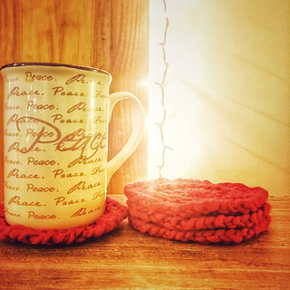 A cup with 'Peace written on it' on a knit coaster next to a stack of three knit coasters. A string of twinkle lights is behind them.