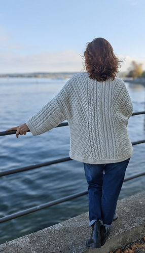 The back showcases elegant design. A striking vertical cable runs down the center, lengthening the silhouette and drawing the eye upward. This architectural centerline is beautifully balanced by diagonal Aran patterns across the shoulders, creating movement and visual interest while framing the back perfectly. At the hemline, a subtle Herringbone pattern provides a grounding finish with soft texture. The interplay of vertical, diagonal, and horizontal stitches creates a harmonious, flattering composition that works beautifully whether worn open or layered.