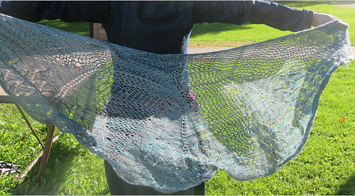 Image Description: a pale blue lace shawl held out behind a person.  It's a half-circle shawl knit in a starburst mesh pattern with a lace pattern stitch at the bottom edge. 