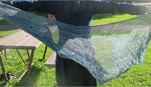 Image Description: a pale blue lace shawl held out behind a person.  It's a half-circle shawl knit in a starburst mesh pattern with a lace pattern stitch at the bottom edge. 
