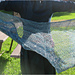 Image Description: a pale blue lace shawl held out behind a person.  It's a half-circle shawl knit in a starburst mesh pattern with a lace pattern stitch at the bottom edge. 