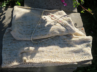 Image Description: A cream skirt laying partly folded on the edge of a garden bed. The skirt is a wrap skirt, knit in stocking stitch with I-cord ties.  5 buttons fasten the front edge of the skirt, and the lower hem is edged with eyelets