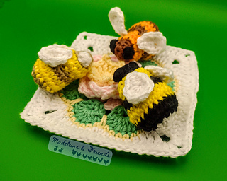 Three crochet bees sitting on a flower themed granny square. The bees are black and yellow, brown and yellow, and brown and orange.