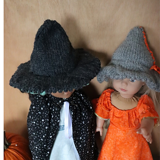 2 dolls side by side, photographed from above. One is wearing a dark grey witches hat with a stiff brim, the other a light grey hat with a floppy brim.