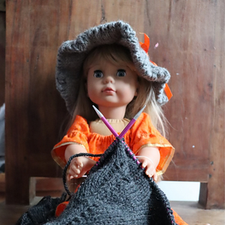 A doll in a grey knitted witches hat and orange dress, sitting. She is holding a dark grey piece of knitting, as if she were knitting.