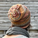A woman with brown curly hair and a grey scarf with her back to the camera with a grey barnboard background wearing a multi-coloured handknit slouchy hat showing the concentric rings on the crown decreases.