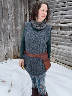 A female is standing facing the camera in front of a barn wall wearing a blue cabled vest and a brown leather belt-pouch.