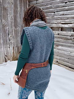 A female is standing facing away from the camera in front of a barn wall wearing a blue cabled vest and a brown leather belt-pouch.