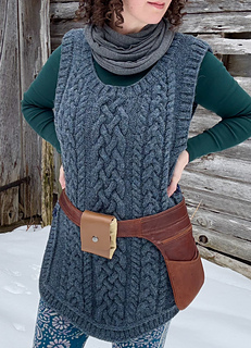 A female is standing with hands on hips facing the camera in front of a barn wall wearing a blue cabled vest and a brown leather belt-pouch.