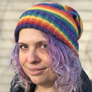 A woman looking at the camera with curly purple hair and wearing a slouchy hat with rainbow stripes.