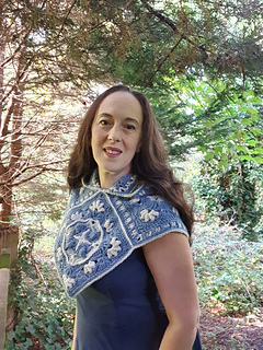 Woman wearing blue and grey crocheted fancy granny square hood in woodland setting (hood down/side-front view)