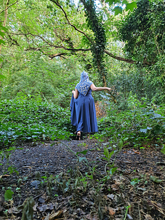 Woman wearing blue and grey crocheted fancy granny square hood in woodland setting (hood up/back view)