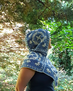 Woman wearing blue and grey crocheted fancy granny square hood in woodland setting (hood up/side view)