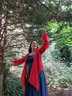 Smiling woman in coral crochet lace wrap cardigan untied  stands in a forest with arms stretched out to the trees