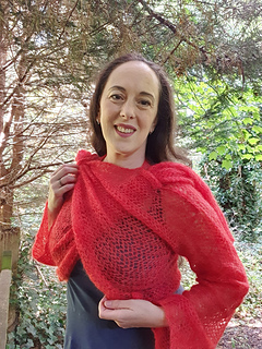 Smiling woman in coral crochet lace wrap cardigan tied asymmetrically at each shoulder stands in a forest holding the shoulder ties