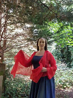 Smiling woman in coral crochet lace wrap cardigan untied  stands in a forest holding the front panels of the cardigan