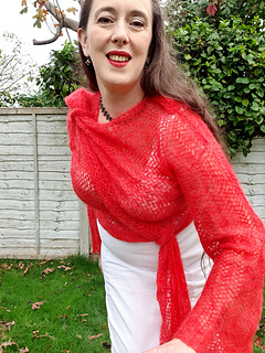 Smiling woman in coral crochet lace wrap cardigan tied asymmetrically at hip and shoulder stands in a garden leaning forwards