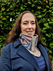 Woman smiling in front of green foliage, wearing a blue coat and a knitted spiral striped cowl.