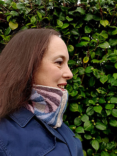 Woman (side profile) in front of green foliage, wearing a blue coat and a knitted spiral striped cowl.