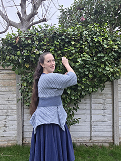 Woman in a blue crocheted tunic poses side on in a garden with green bushes and bare tree. Wooden fence in the background. Calm expression.