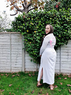 Woman in a white knitted shrug with ribbon bows smiles outdoors. Greenery and a wooden fence are in the background on a sunny day.