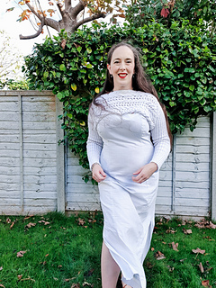 Woman in a white knitted cropped pullover smiles outdoors. Greenery and a wooden fence are in the background on a sunny day.