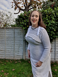 Woman in a white knitted cropped pullover with ribbon bows smiles outdoors. Greenery and a wooden fence are in the background on a sunny day.