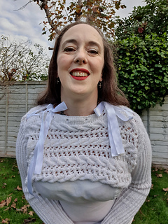 Woman in a white knitted cropped pullover with ribbon bows smiles outdoors. Greenery and a wooden fence are in the background on a sunny day.