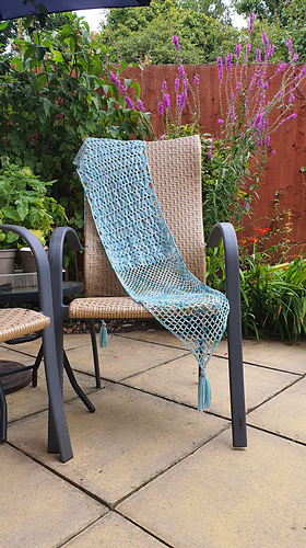 light blue variegated hairpin crocheted shawl on a chair in a garden. Greenery and a red fence are in the background.