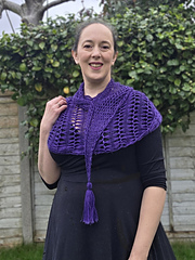 Woman in a black dress with a purple variegated hairpin crocheted shawl stands in a garden. Greenery and a fence are in the background.