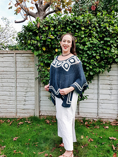 Woman smiling in a garden wearing a blue and white poncho. Wooden fence and leafless trees in the background under a cloudy sky.