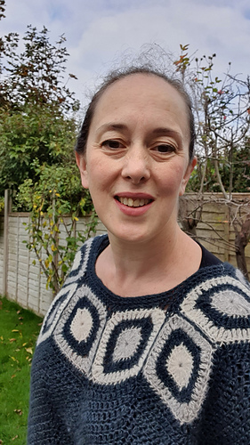 Woman smiling in a garden wearing a blue and white poncho. Wooden fence and leafless trees in the background under a cloudy sky.