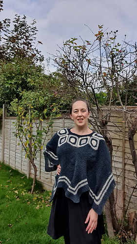 Woman smiling in a garden wearing a blue and white poncho. Wooden fence and leafless trees in the background under a cloudy sky.
