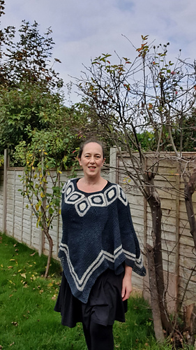Woman smiling in a garden wearing a blue and white poncho. Wooden fence and leafless trees in the background under a cloudy sky.